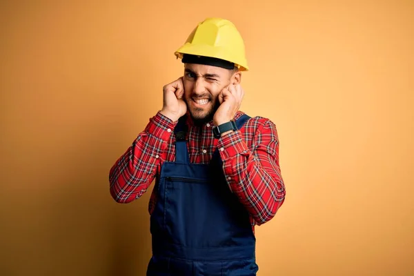 年轻的建筑工人身穿建筑制服 头戴安全帽 戴黄色的隔离背景 用手指捂住耳朵 对嘈杂的音乐感到恼怒 聋人概念 — 图库照片