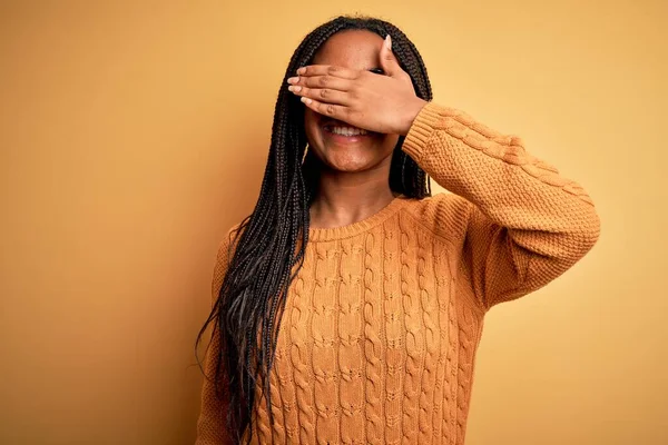 Young african american smart woman wearing glasses and casual sweater over yellow background smiling and laughing with hand on face covering eyes for surprise. Blind concept.