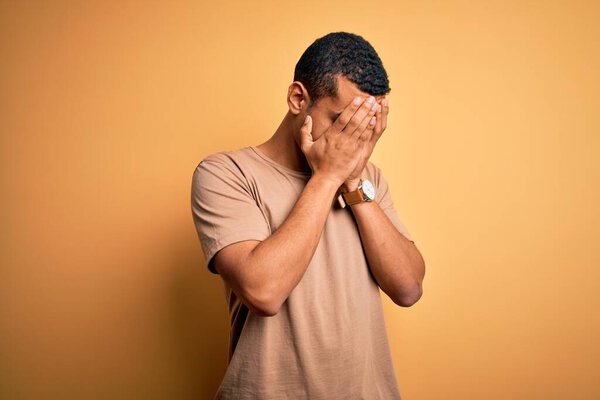 Young handsome african american man wearing casual t-shirt standing over yellow background with sad expression covering face with hands while crying. Depression concept.