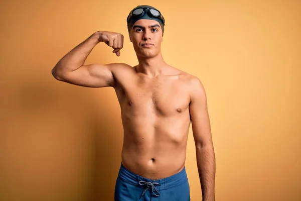 年轻英俊的男子赤身裸体 身穿泳衣 头戴泳帽 身披孤立的黄色背景 强壮的男子展示臂膀肌肉 自信而自豪 — 图库照片
