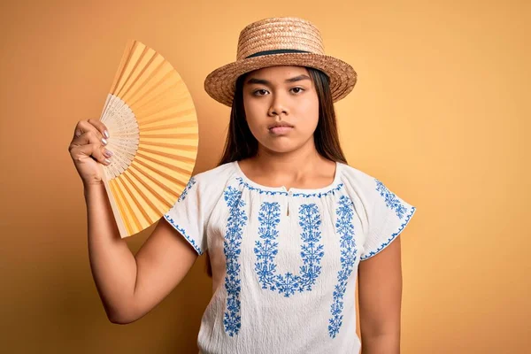 年轻的亚洲女游客在度假时戴着夏帽 手扇罩在黄色的背景上 脸上带着自信的表情 脸上带着严肃的表情 — 图库照片