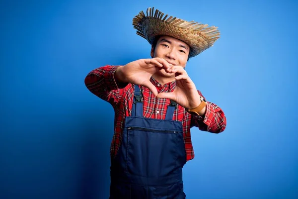 Young handsome chinese farmer man wearing apron and straw hat over blue background smiling in love doing heart symbol shape with hands. Romantic concept.