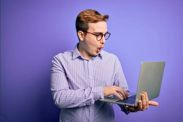 年轻英俊的红头发商人在紫色背景的笔记本电脑上工作时 吓得满脸诧异 吓得面无血色 吓得面无血色 — 图库照片
