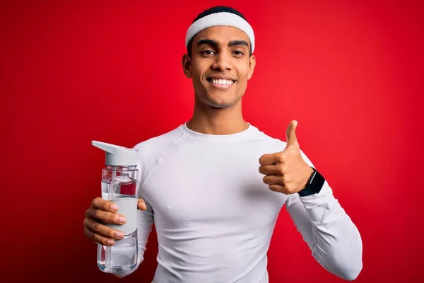 Young handsome african american sportsman wearing sportswear drinking bottle of water happy with big smile doing ok sign, thumb up with fingers, excellent sign