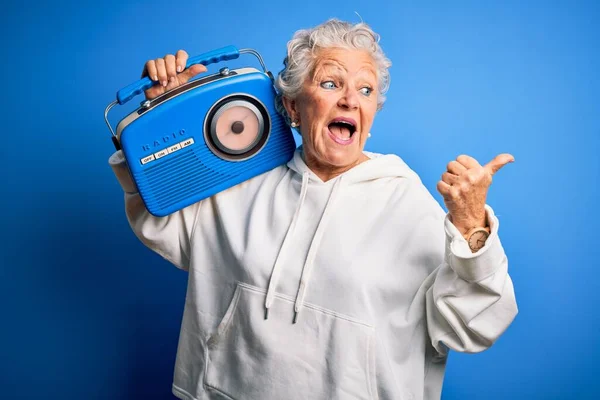 年长的漂亮女人拿着老式收音机 站在孤立的蓝色背景上 用拇指指向旁边 笑容满面 — 图库照片