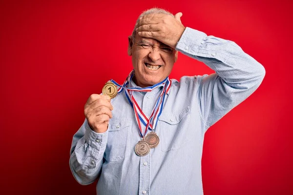 Middle age hoary champion man wearing medals standing over isolated red background stressed with hand on head, shocked with shame and surprise face, angry and frustrated. Fear and upset for mistake.