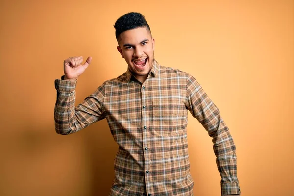 Genç yakışıklı adam, günlük gömleğini giymiş, izole edilmiş sarı arka planda duruyor mutlu ve neşeli bir şekilde dans ediyor, hareketli ve kendinden emin bir şekilde müzik dinliyor.