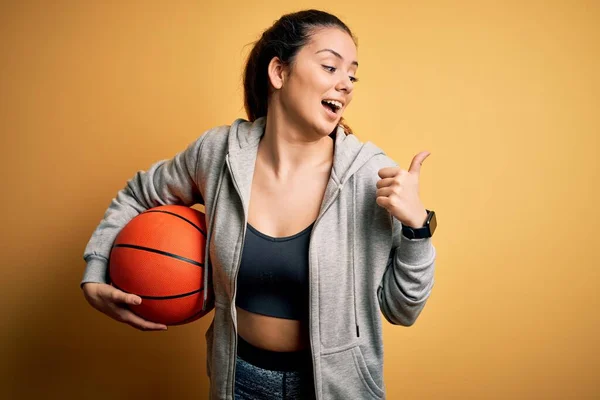 年轻美丽的黑发女运动员 背景是黄色的 手握篮球 手握大拇指 笑容满面地站在旁边 — 图库照片