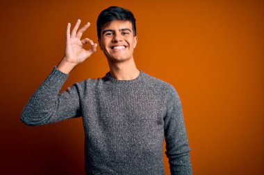 İzole edilmiş turuncu arka plan üzerinde duran sıradan süveter giyen yakışıklı genç adam el ve parmaklarıyla 
