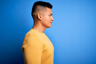 Sarı günlük süveter giyen genç, yakışıklı, latin bir adam. Mavi arka plan üzerinde. Yan tarafa bakıyor, rahat profil pozu veriyor..