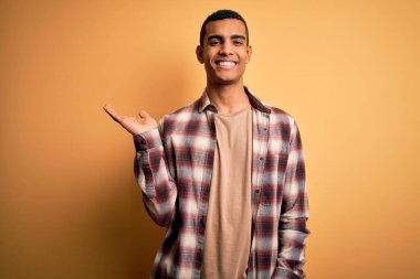 Genç, yakışıklı, Afro-Amerikalı bir adam sarı arka planda dikilmiş gülümsüyor ve avucunun içi kameraya bakıyor..