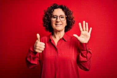 Orta yaşlı, kıvırcık saçlı, rahat gömlekli, kırmızı arka planda gözlüklü güzel bir kadın. Altıncı parmaklarıyla işaret ederken kendinden emin ve mutlu bir şekilde gülümsüyor..