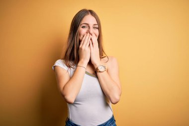 Genç, güzel, kızıl saçlı bir kadın izole edilmiş sarı arka planda günlük tişört giyip gülüyor ve kıkırdıyor. Ağzı ellerle, dedikoduyla ve skandalla örtüyor.