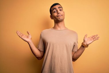 Sarı arka plan üzerinde sıradan tişörtler giyen, elleri ve kolları havada, kafası karışmış genç, Afro-Amerikan bir adam. Şüphe kavramı.