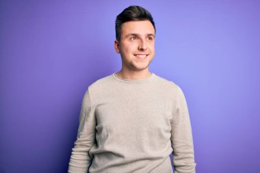 Mor yalıtılmış arka plan üzerine günlük süveter giyen yakışıklı beyaz bir adam yüzünde gülümsemeyle, doğal bir ifadeyle yan yana bakıyor. Kendinden emin gülen.