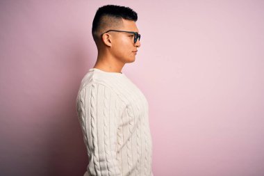 Beyaz süveter giyen ve pembe arka planda gözlüklü genç, yakışıklı Latin bir adam. Yan tarafa bakıyor, rahat profil pozu veriyor, kendine güvenen bir gülümsemesi var..