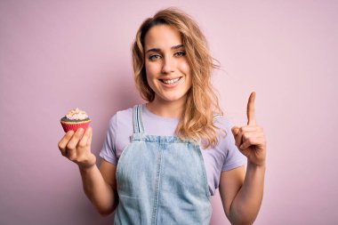 Genç, güzel, sarışın bir kadın izole edilmiş pembe arka planda çikolatalı kek. Bir fikir ya da soru işareti ile şaşırmış, mutlu yüzlü, bir numara.
