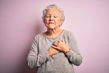 Üst düzey güzel bir kadın, günlük tişört giyer, pembe arka plan üzerinde durur, elleri göğsünde, gözleri kapalı ve yüzünde minnet dolu bir jest ile gülümser. Sağlık kavramı.