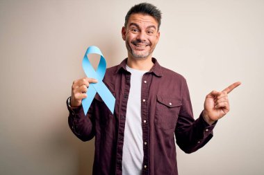İzole edilmiş beyaz arka planda mavi kanser kurdelesi tutan genç yakışıklı bir adam çok mutlu bir şekilde elini ve parmağını kenarı işaret ediyor.
