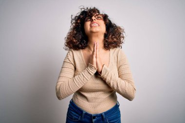 Genç ve güzel kıvırcık Arap kadın, beyaz arka planda günlük tişört ve gözlük takıyor, yalvarıyor ve dua ediyor. Yüzünde umut ifadesi olan çok duygusal ve endişeli bir ifade var. Yalvarıyorum..