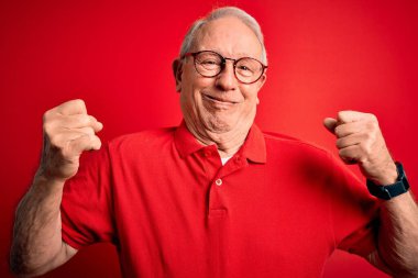 Kırmızı arka planda gri saçlı, gözlüklü ve rahat tişörtlü yaşlı adam gururla gülümseyen kol kaslarını gösteriyor. Fitness konsepti.