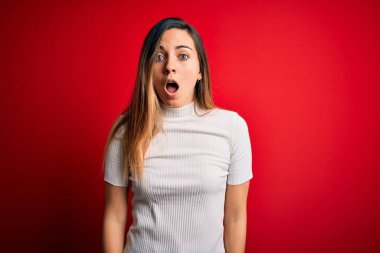 Güzel sarışın kadın, mavi gözlü, kırmızı arka planda sıradan beyaz tişörtler giyiyor. Şaşırmış ve şaşırmış bir ifade, korku ve heyecanlı bir yüz ifadesi var..