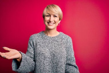 Modern kısa saçlı genç bir kadın pembe arka planda rahat bir süveter giyiyor. Gülümsüyor, kollarını açıp gülümsüyor. Dostça karşılıyor, olumlu ve kendinden emin selamlıyor.