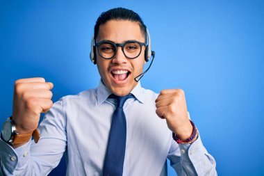 Gözlüklü ve kravatlı genç Brezilyalı çağrı merkezi ajanı kolları açık ve kolları açık bir şekilde başarıyı kutlayan kulaklıkla çalışıyor. Kazanan konsept.
