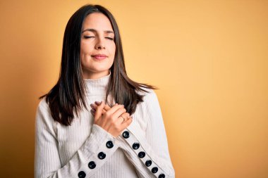 Mavi gözlü genç esmer kadın izole edilmiş sarı arka planda günlük süveter giyiyor elleri göğsünde, gözleri kapalı ve yüzünde minnet dolu bir jest ile gülümsüyor. Sağlık kavramı.