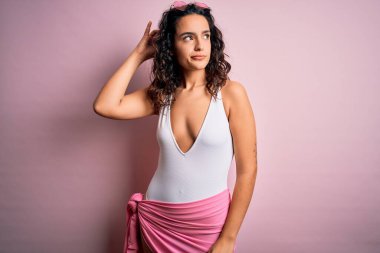 Tatilde kıvırcık saçlı, pembe arka planda beyaz mayo giyen, kafası karışık ve soru soran güzel bir kadın. Şüpheyle kararsız, kafa kafaya düşünerek. Düşünceli kavram.