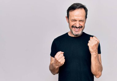 Sıradan siyah tişört giyen orta yaşlı bir adam izole edilmiş beyaz arka planda dikilmiş şaşırmış ve şaşırmış bir şekilde kolları havada ve gözleri kapalı bir şekilde başarıyı kutluyor.