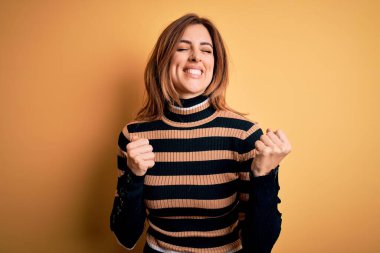 Sarı arka plan üzerine çizgili balıkçı yaka kazak giyen genç ve güzel bir kadın. Kolları havada ve gözleri kapalı zaferi kutlarken gülümsüyor. Kazanan konsept.