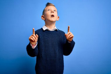 Mavi arka planda mavi süveter giyen küçük beyaz çocuk şaşırmış ve şaşırmış görünüyor ve parmaklarıyla işaret ediyor ve kollarını kaldırıyor..