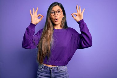 Mor izole edilmiş arka planda gözlük takan güzel ve zeki bir kadın. Şaşırmış ve şaşırmış görünüyor. Parmaklarıyla onay sembolü yapıyor. Çılgın ifade