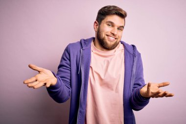 Sarışın, sakallı ve mavi gözlü pembe arka planda mor eşofman üstü giyen, elleri ve kolları havada, kafası karışmış genç bir adam. Şüphe kavramı.
