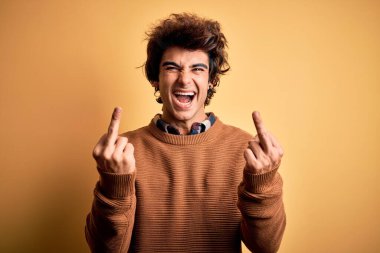 Young handsome man wearing casual shirt and sweater over isolated yellow background Showing middle finger doing fuck you bad expression, provocation and rude attitude. Screaming excited