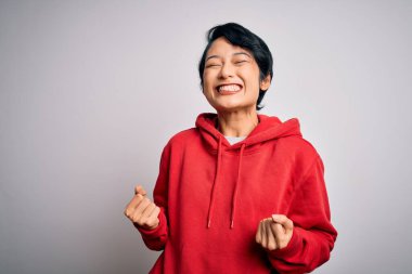 Genç, güzel Asyalı bir kız beyaz arka planda kapüşonlu sıradan bir sweatshirt giyiyor çok mutlu ve heyecanlı bir şekilde kollarını kaldırıp, gülümseyip başarı için bağırıyor. Kutlama konsepti.