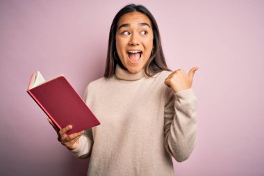 Genç Asyalı akıllı kadın kitap günlüğü tutuyor pembe arka plan üzerinde duruyor ve gülümseyen bir gülümsemeyle baş parmağıyla işaret ediyor.