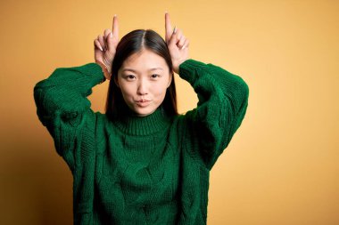 Sarı arka plan üzerine yeşil kazak giyen genç, güzel Asyalı kadın boynuzları gibi parmağıyla komik hareketler yapıyor.