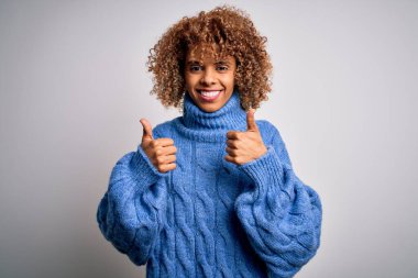 Beyaz arka plan işareti üzerine balıkçı yaka kazak giyen güzel Afro-Amerikan kadın. Elleriyle olumlu jest yapıyor. Gülümsüyor ve mutlu. Neşeli ifade ve kazanan jest.