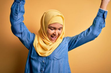 Kıvırcık saçlı genç bir kadın sarı arka planda geleneksel Arap başörtüsü takıyor mutlu ve neşeli dans ediyor, hareketli, rahat ve kendinden emin bir şekilde müzik dinliyor.