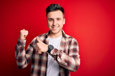 Kırmızı izole arka plan üzerine gündelik modern gömlek giyen genç, beyaz bir adam. Arka tarafı işaret ediyor. El ve baş parmakları havada, kendine güvenen.