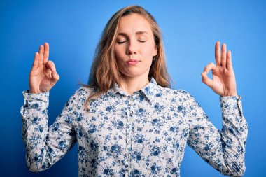 Mavi arka planda dikilen sıradan bir gömlek giyen genç, güzel sarışın kadın rahatla ve parmaklarıyla meditasyon hareketi yaparken gözleri kapalı gülümse. Yoga konsepti.