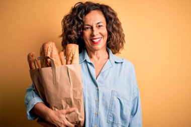 Orta yaşlı, güzel bir kadın, elinde kağıt torbayla izole edilmiş sarı arka planda, mutlu bir yüzü olan, kendine güvenen bir gülümsemeyle, dişlerini gösteren bir çanta tutuyor.