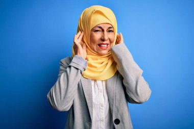 Orta yaşlı esmer iş kadını, geleneksel Müslüman tesettüre bürünmüş mavi arka planda, yüksek sesli müzik sesi için parmaklarıyla kulaklarını kapatıyor. Sağır kavramı.