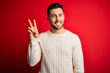 İzole edilmiş kırmızı arka plan üzerinde duran, rahat beyaz süveter giyen genç yakışıklı adam iki numaralı parmaklarıyla işaret ederken kendinden emin ve mutlu bir şekilde gülümsüyor..