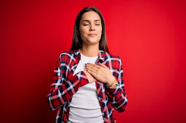 Genç, güzel, esmer, günlük gömlekler giyen, kırmızı arka planda duran, elleri göğsünde, gözleri kapalı ve yüzünde minnet dolu bir jest yapan bir kadın. Sağlık kavramı.