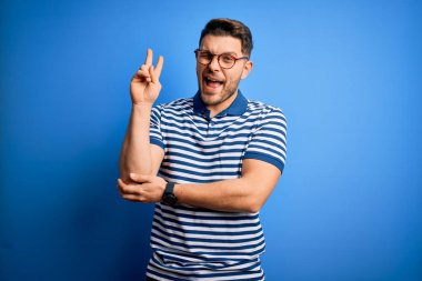 Mavi gözlü, gözlüklü ve mavi arka planda sıradan çizgili tişörtlü mutlu yüzüyle kameraya bakıp zafer işareti yapan genç bir adam. İki numara..