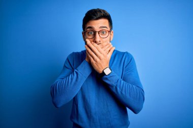 Sakallı, sıradan süveter ve mavi arka planda gözlük takan genç yakışıklı bir adam yanlışlıkla ağızlarını kapatarak şok geçirdi. Gizli konsept.