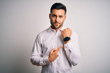 Zarif gömlekli genç adam izole edilmiş beyaz arka planda dikiliyor. Aceleyle saati, sabırsızlığı ve rahat bir ifadeyle kameraya bakıyor.